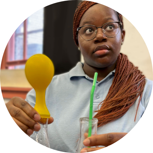 A person in glasses holds a yellow balloon over a glass container with liquid, while also holding a green straw.