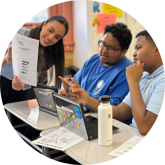 A teacher stands by two students seated at desks, showing them a paper while they work on their laptops. The students are engaged and focused, with one holding a pen and the other wearing glasses.