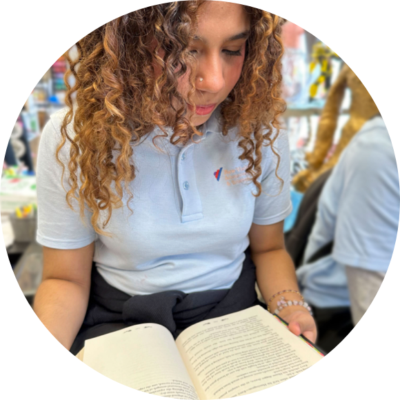 Young woman with curly hair reading a book while wearing a light blue polo shirt, seated in what appears to be a casual setting.