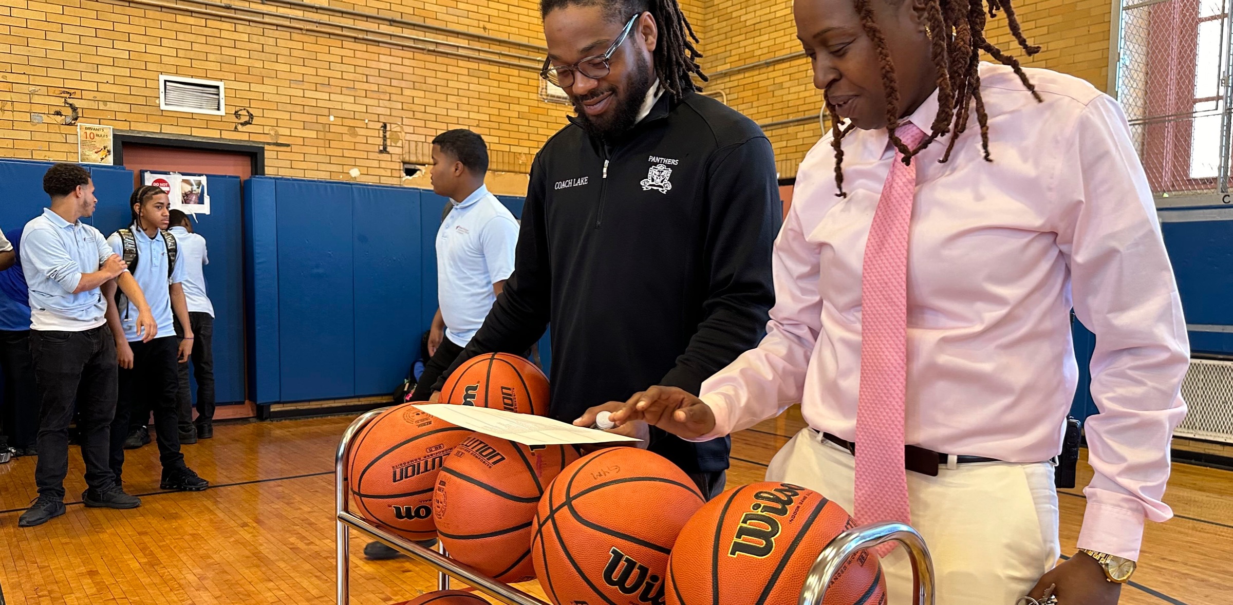 Two men in a gymnasium inspect documents on a cart holding several basketballs. People are seen conversing in the background.