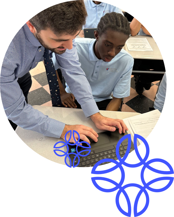 An instructor in a striped shirt assists a student with a braided hairstyle on a laptop at a table, while other students work in the background.