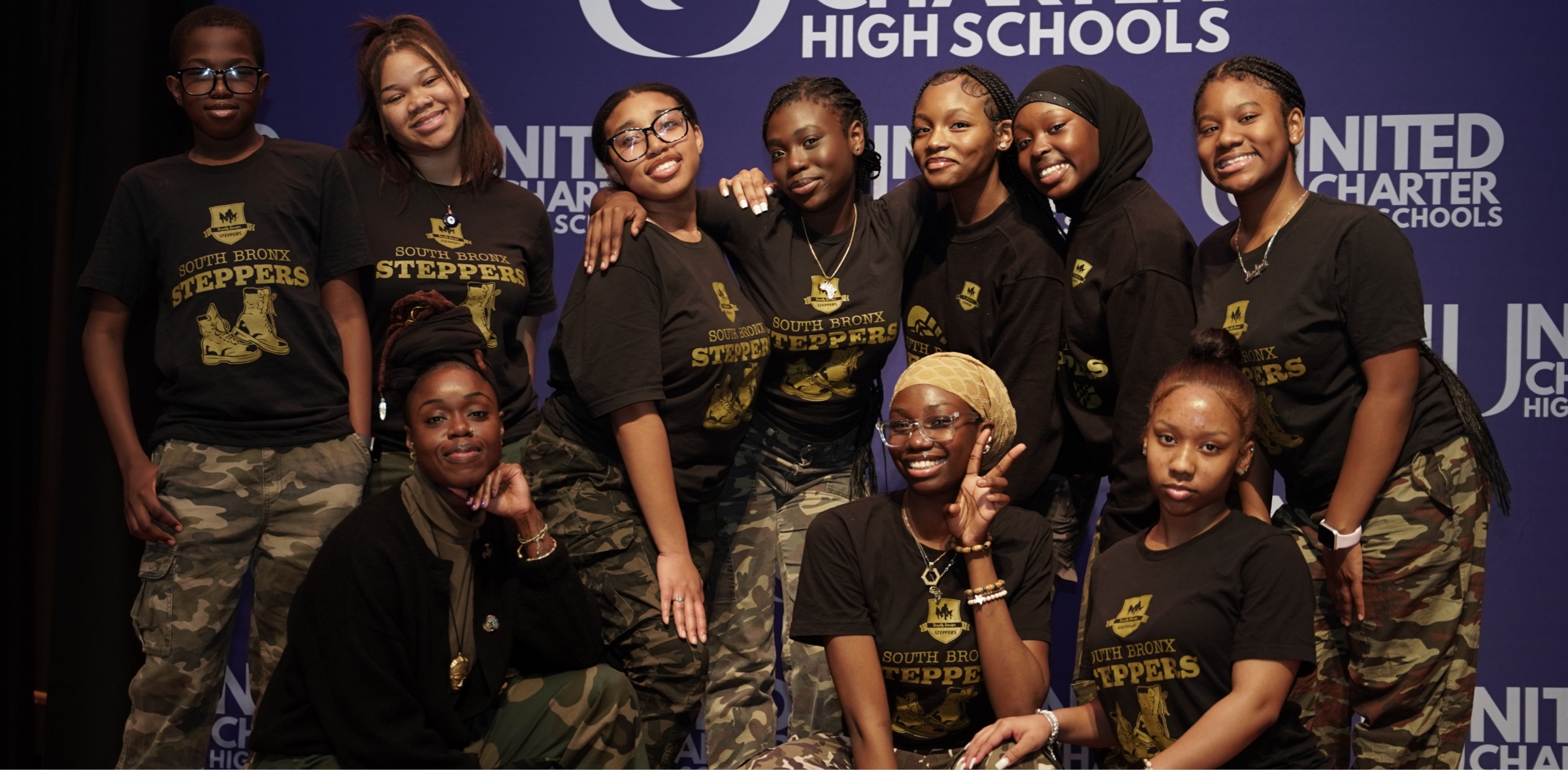 A group of young individuals posing on stage, wearing matching "South Bronx Steppers" t-shirts and camo pants, with the backdrop displaying "United Charter High Schools.