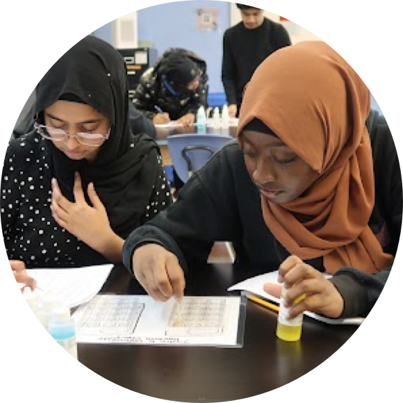 Two students wearing hijabs work on a science experiment with worksheets and test tubes in a classroom setting. Other students are visible in the background.