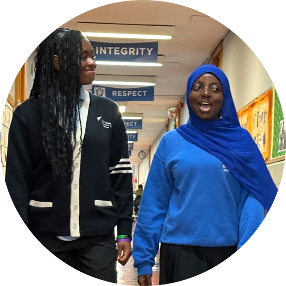 Two students in school uniforms walking down a hallway adorned with banners that read "INTEGRITY" and "RESPECT." One wears a cardigan and the other a blue hijab.