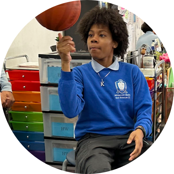 A person in a blue sweatshirt spins a basketball on their finger while sitting in a classroom with colorful drawers in the background.