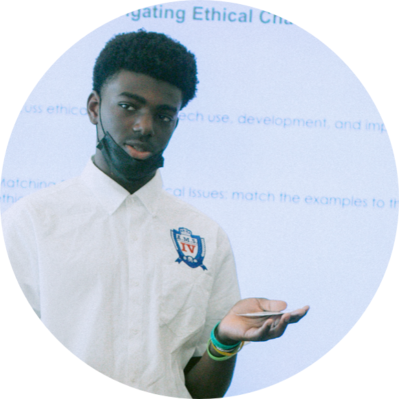 A person wearing a white shirt with an emblem, holding a pen, and standing in front of a screen displaying text.