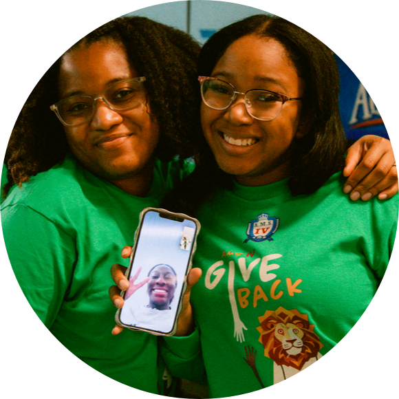 Two people wearing green shirts and glasses smile while holding a phone displaying a video call with another smiling person. The shirts have a graphic of a lion and the text "GIVE BACK.