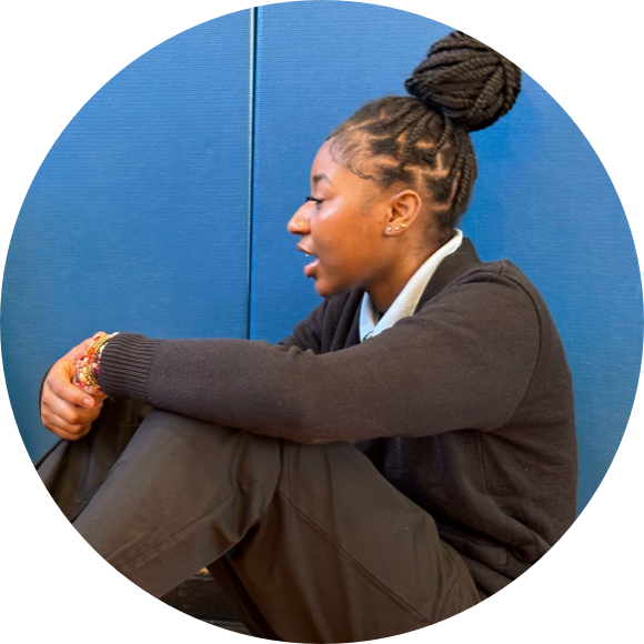 A person with braided hair sits against a blue wall, wearing a dark sweater and pants, speaking while resting their elbows on their knees.