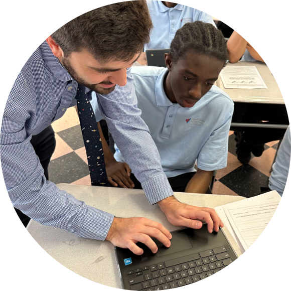 A teacher in a checkered shirt assists a student with a braided hairstyle, who is wearing a blue polo shirt, as they work on a laptop in a classroom.