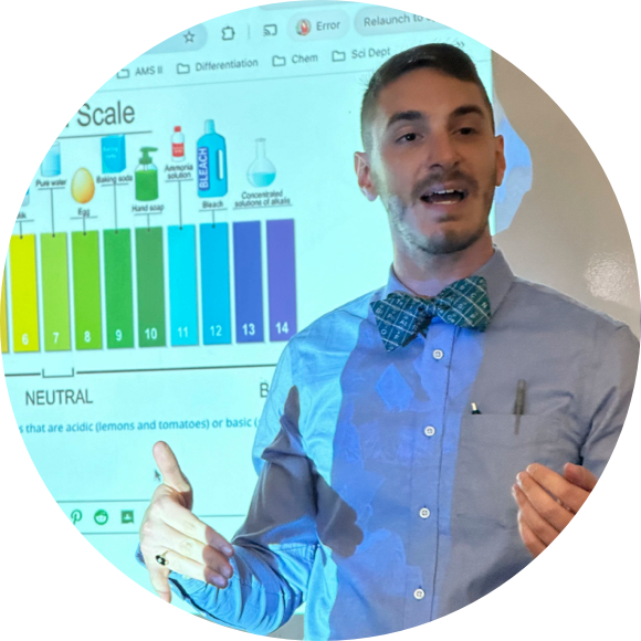 A person is speaking in front of a projected image showing a pH scale diagram. They are wearing a shirt, bow tie, and have a pen clipped to their pocket.