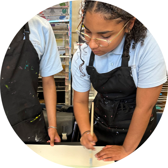 Two individuals wearing black aprons and light blue shirts are painting. One person is focused on painting while the other points at something on the table.