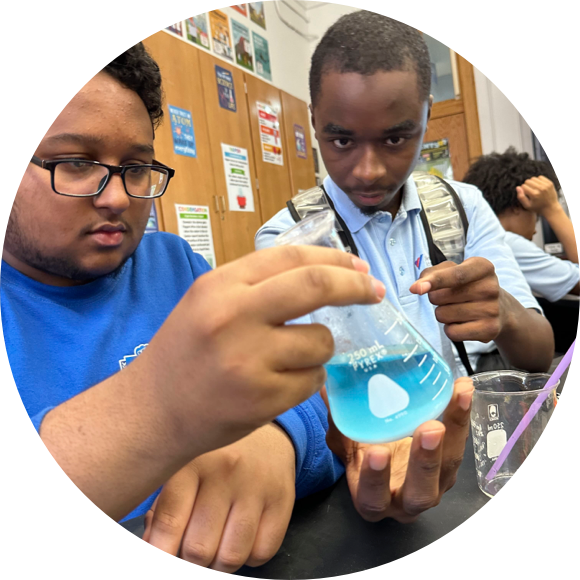 Two students in a classroom look intently at a beaker filled with blue liquid. One student is wearing glasses and a blue shirt, while the other has a serious expression and is holding the beaker.