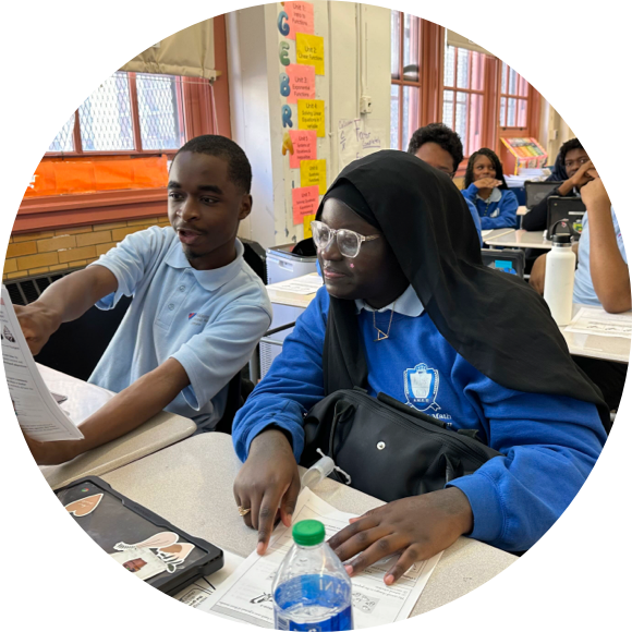 Two students in a classroom are looking at a paper and discussing it. Both are seated at desks with worksheets and water bottles in front of them. Other students are visible in the background.