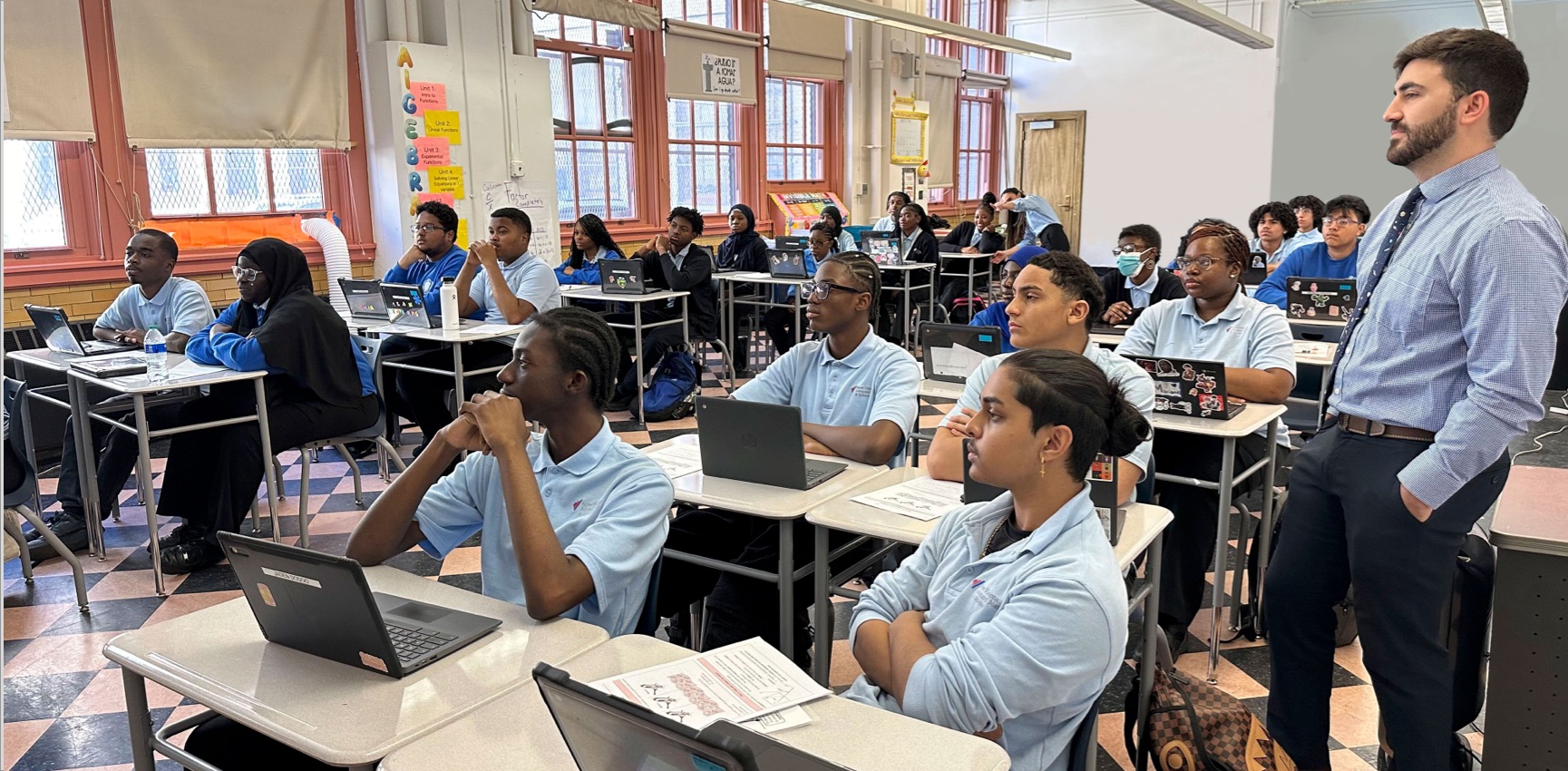 A classroom with students in uniforms and a teacher standing, all engaged in looking towards the front. Desks have laptops and papers on them.