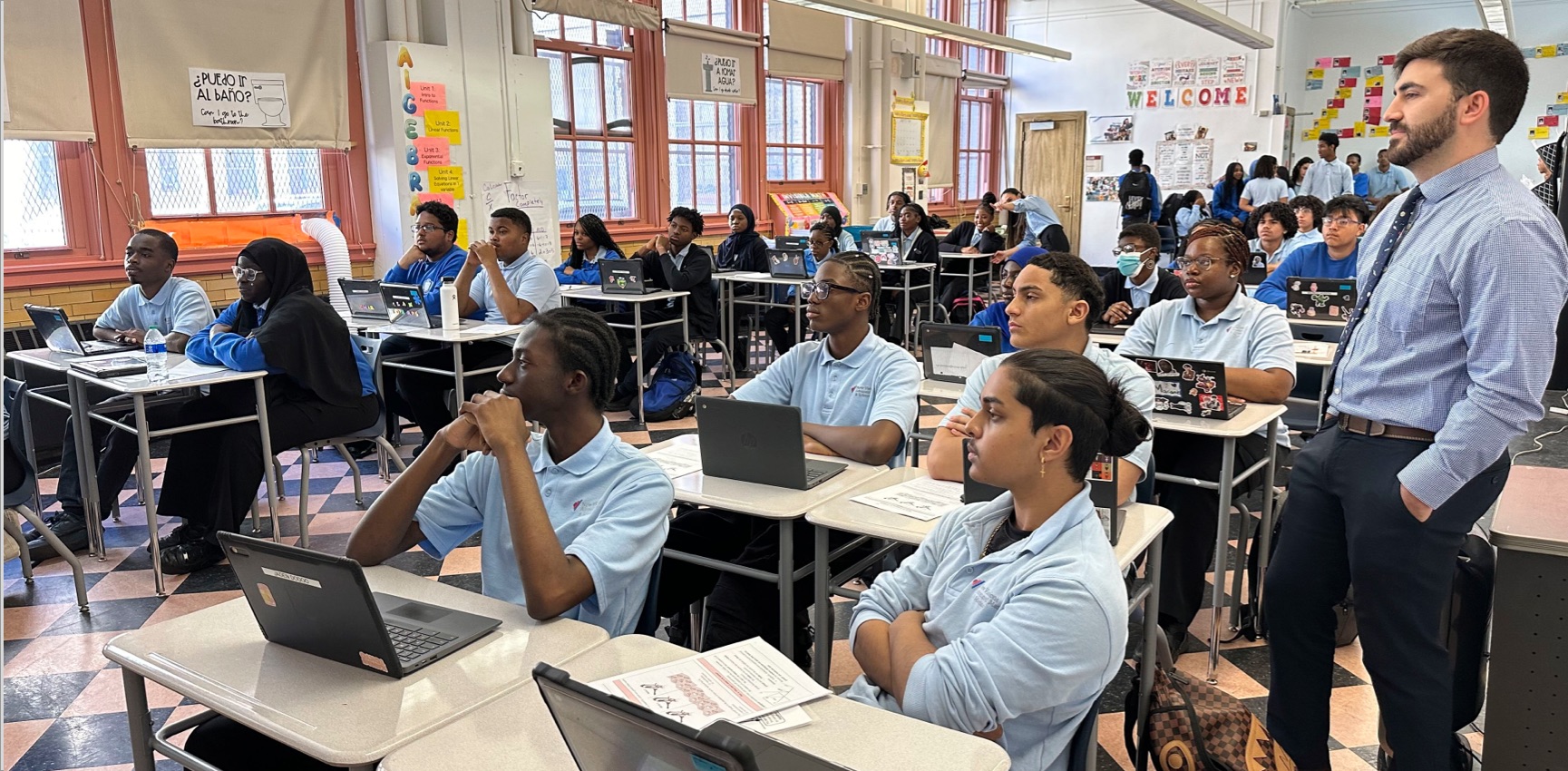 Students in uniforms sit at desks with laptops in a classroom, engaged in a lesson. A teacher stands nearby, overseeing the students. Classroom decorations and posters are visible.