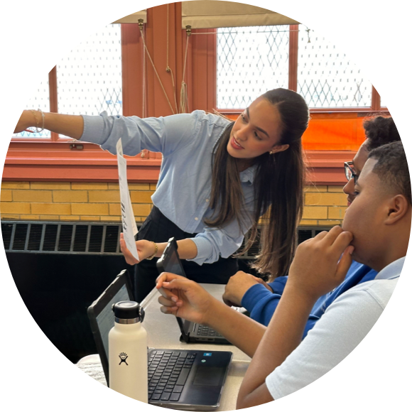 A woman in a light blue shirt explains a document to two seated individuals using laptops in a classroom setting.