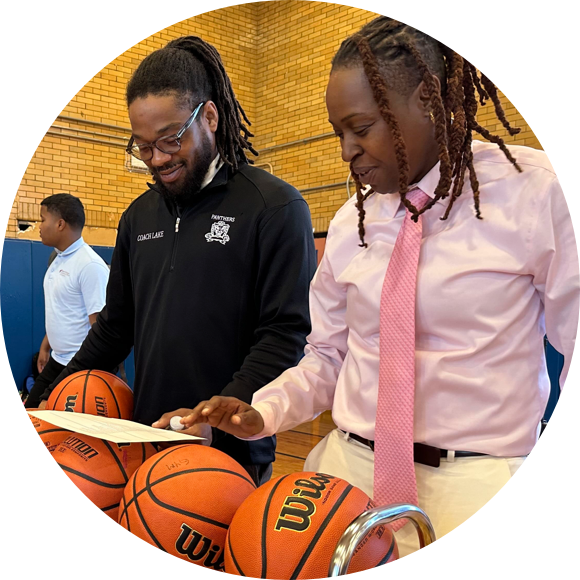 Two people stand near a rack of basketballs in a gym, looking at a document. One wears a black jacket and the other a pink shirt and tie. A third person is in the background.