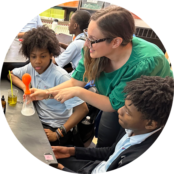 A teacher in a green shirt demonstrates a science experiment to two attentive students. A balloon inflates over a beaker containing white foam.