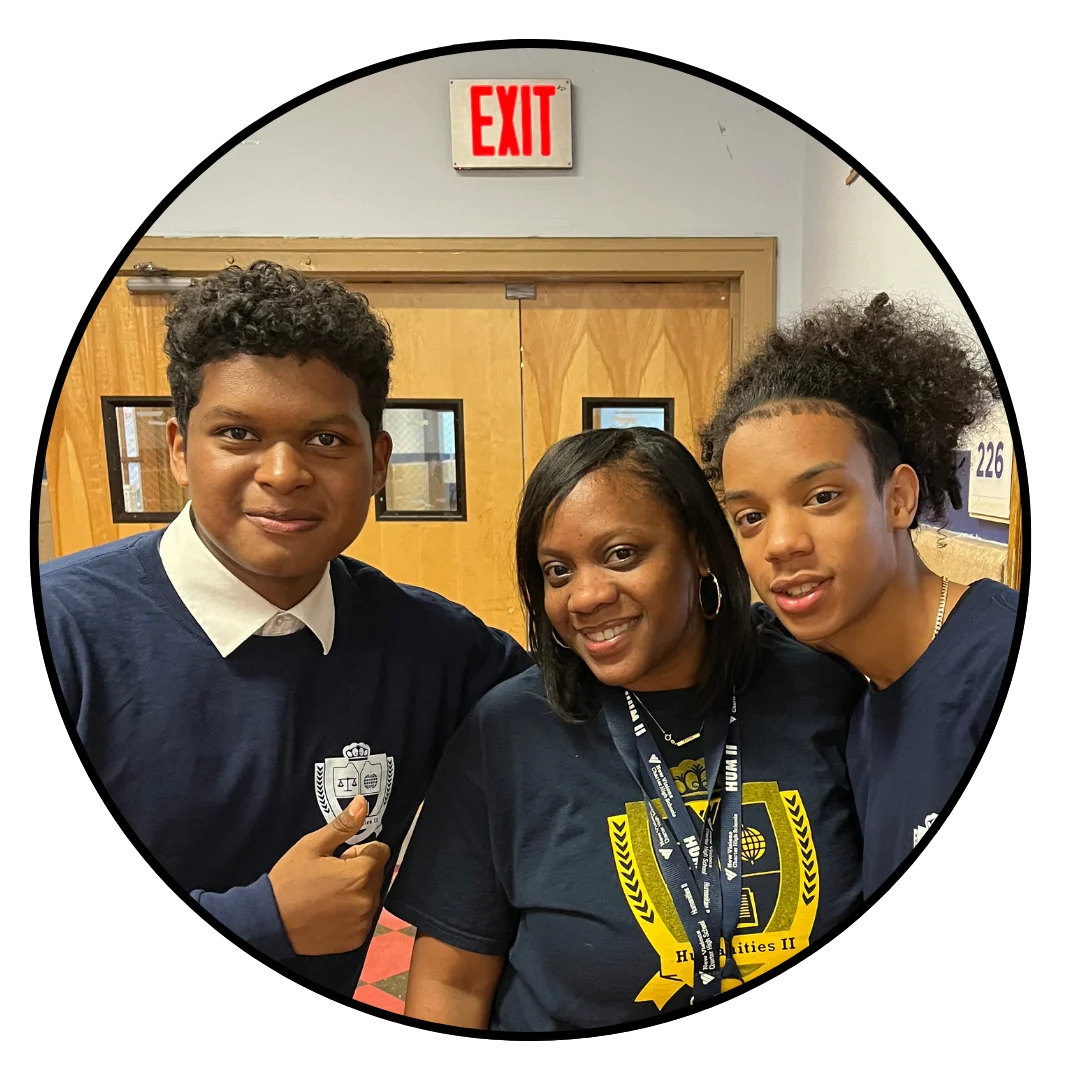 Three people standing together indoors, smiling at the camera. The woman in the center is wearing a lanyard, and the two young men are wearing similar navy blue tops. An exit sign is visible above.