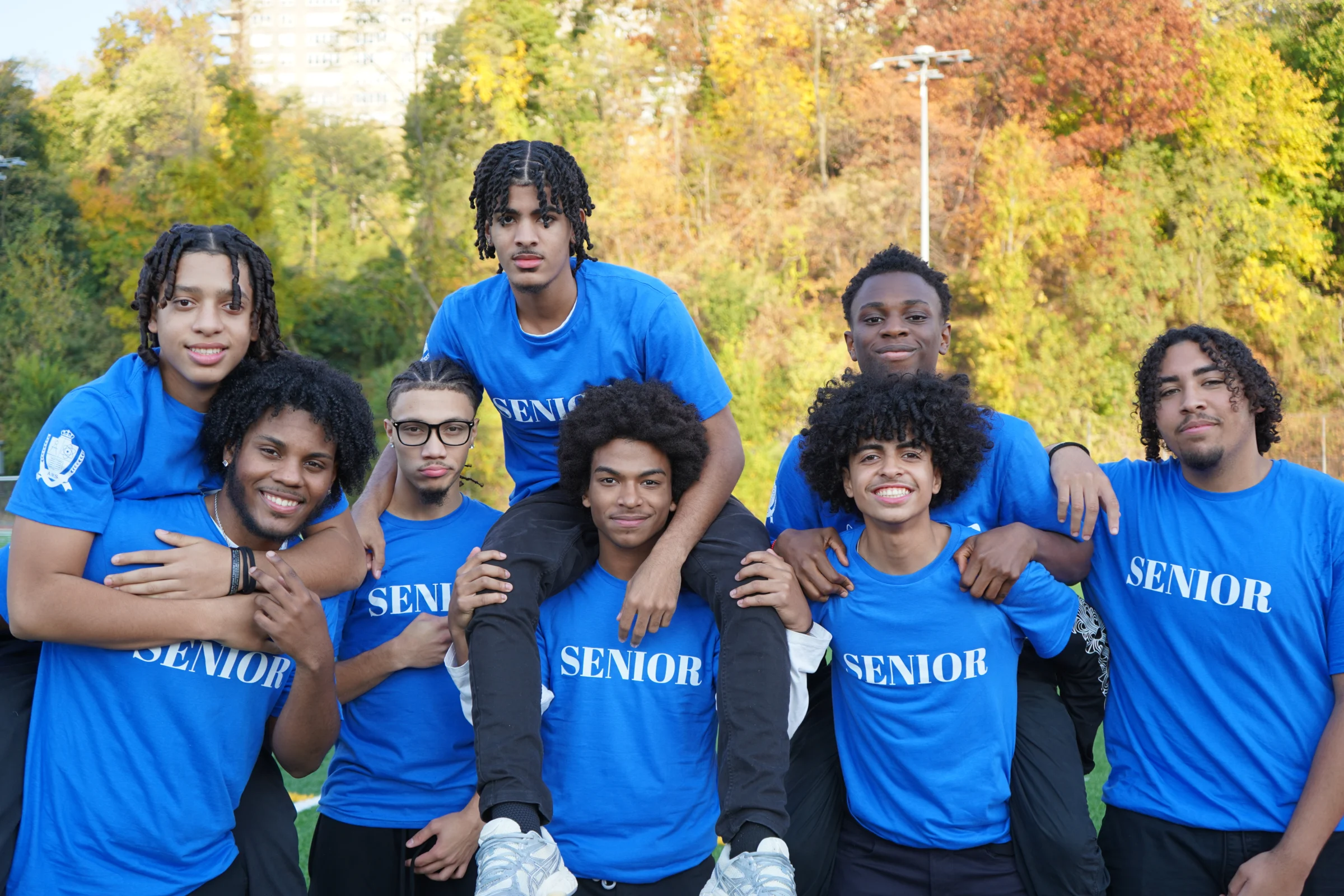 A group of eight young people wearing blue "Senior" shirts pose outdoors, with trees and a building in the background.