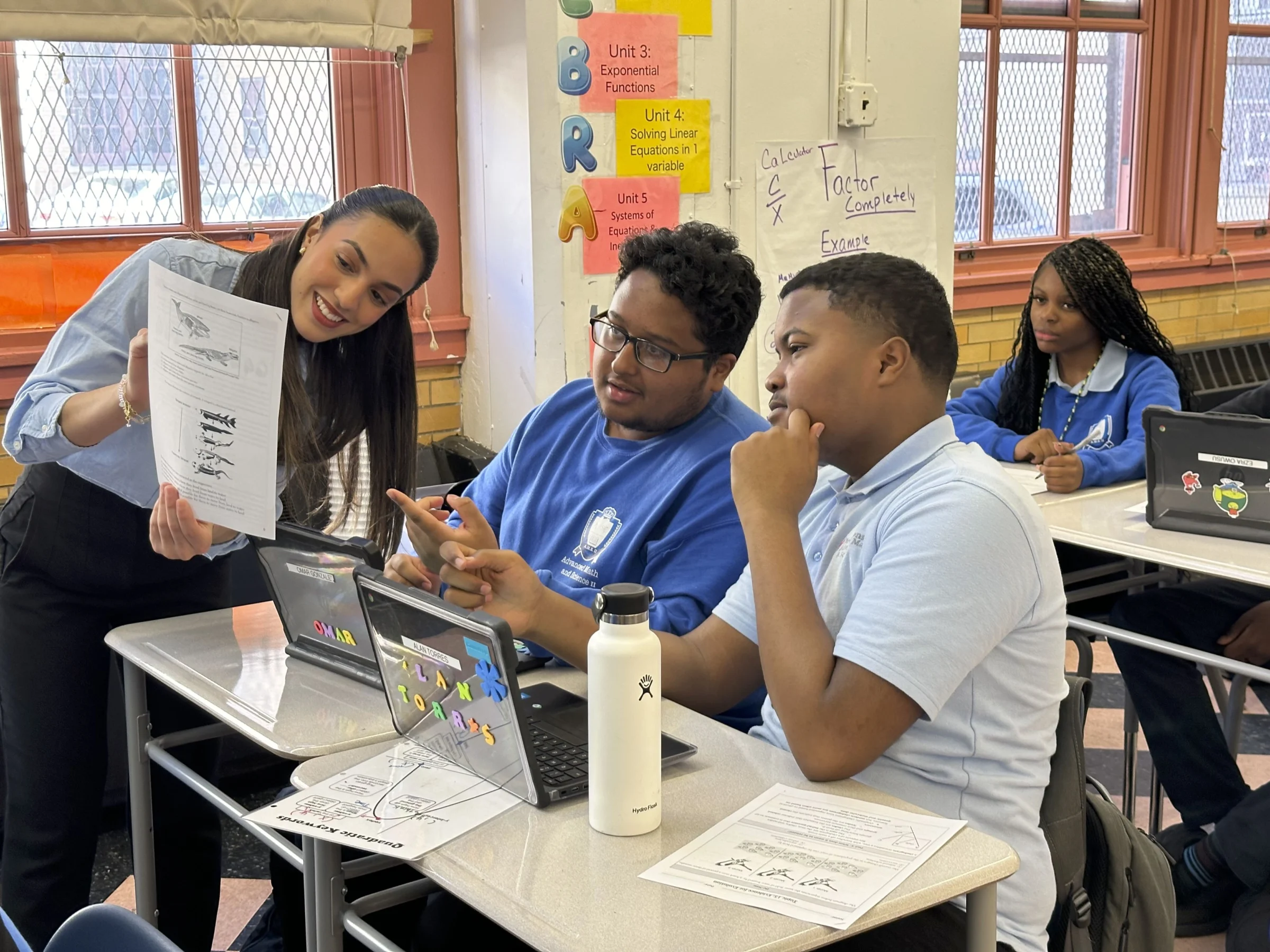 A teacher shows a paper to two engaged students at their desks, with laptops open. Bright classroom with educational posters on the walls.