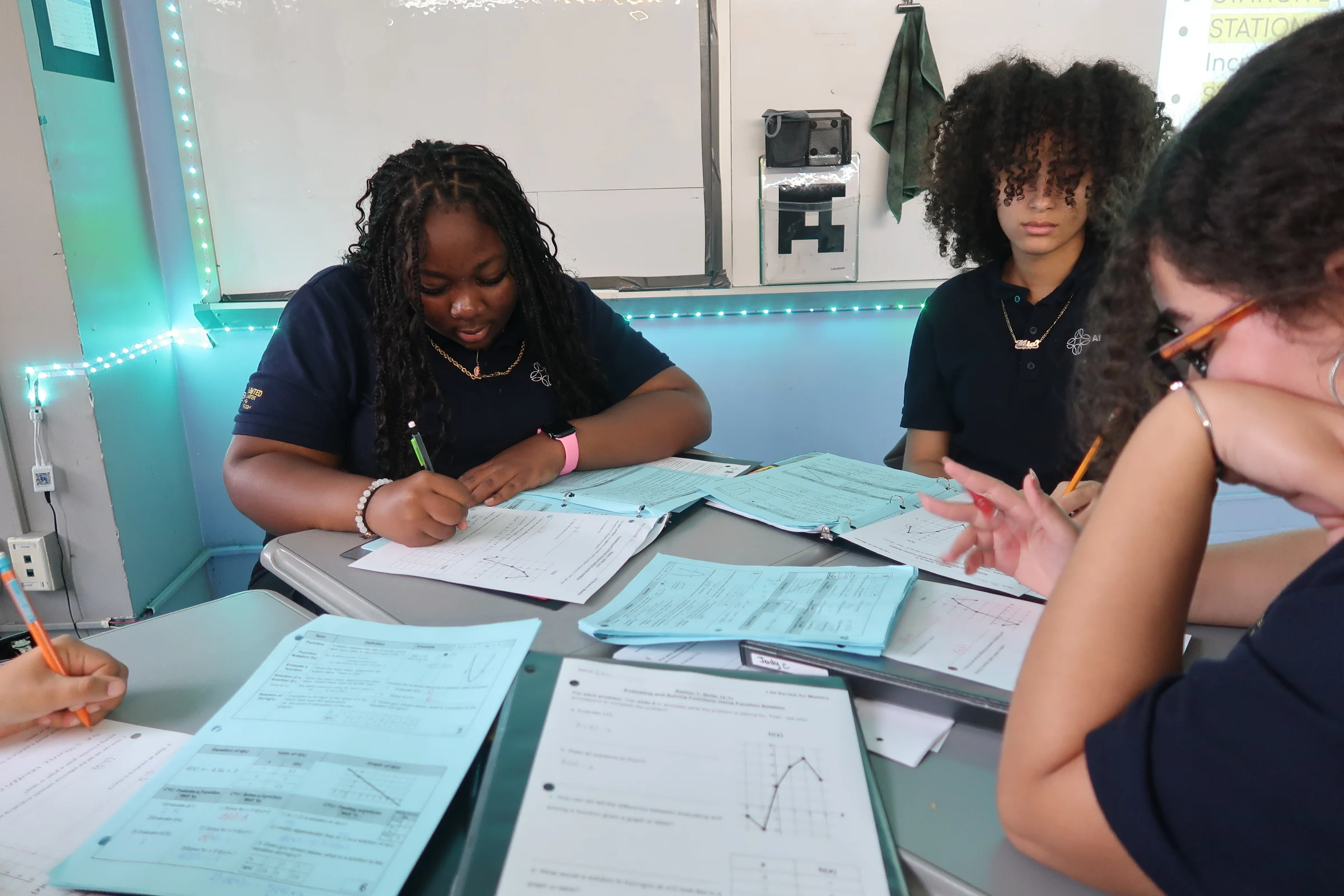 Students in a classroom are focused on writing and studying worksheets at a shared desk.