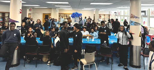 A large group of people gather in a room decorated with banners and blue tablecloths. Some are sitting and others standing, with one person holding blue balloons in the background.