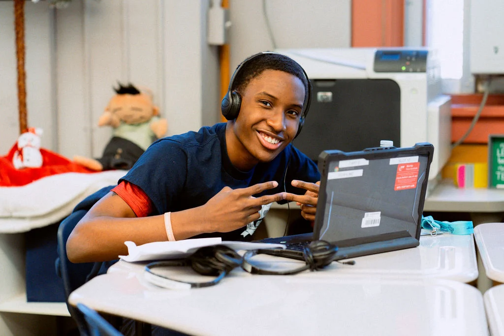 A person smiling and gesturing peace signs while seated at a desk with a laptop and headphones in a classroom setting.