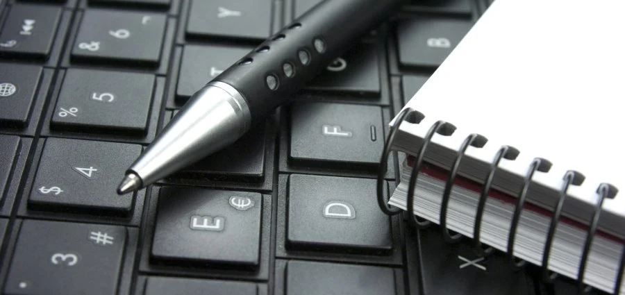 A pen and a spiral notebook are placed on a black computer keyboard.