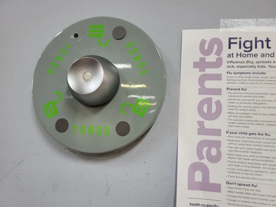 A Yondr phone pouch station mounted on a wall next to a health information poster titled "Parents: Fight Flu at Home and...".