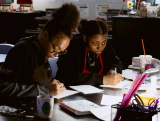 Two students sitting at a desk in a classroom, focused on writing tasks. Various school supplies are scattered on the table.