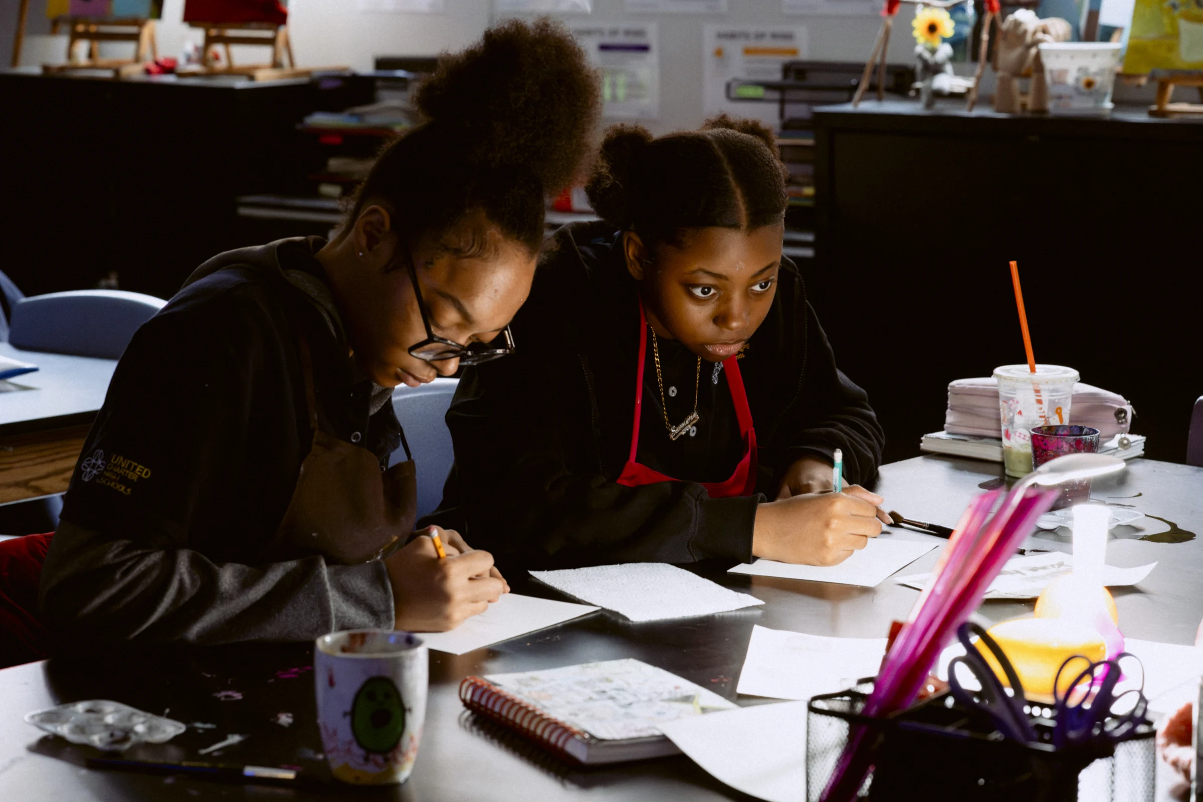 Two students sitting at a desk in a classroom, focused on writing tasks. Various school supplies are scattered on the table.