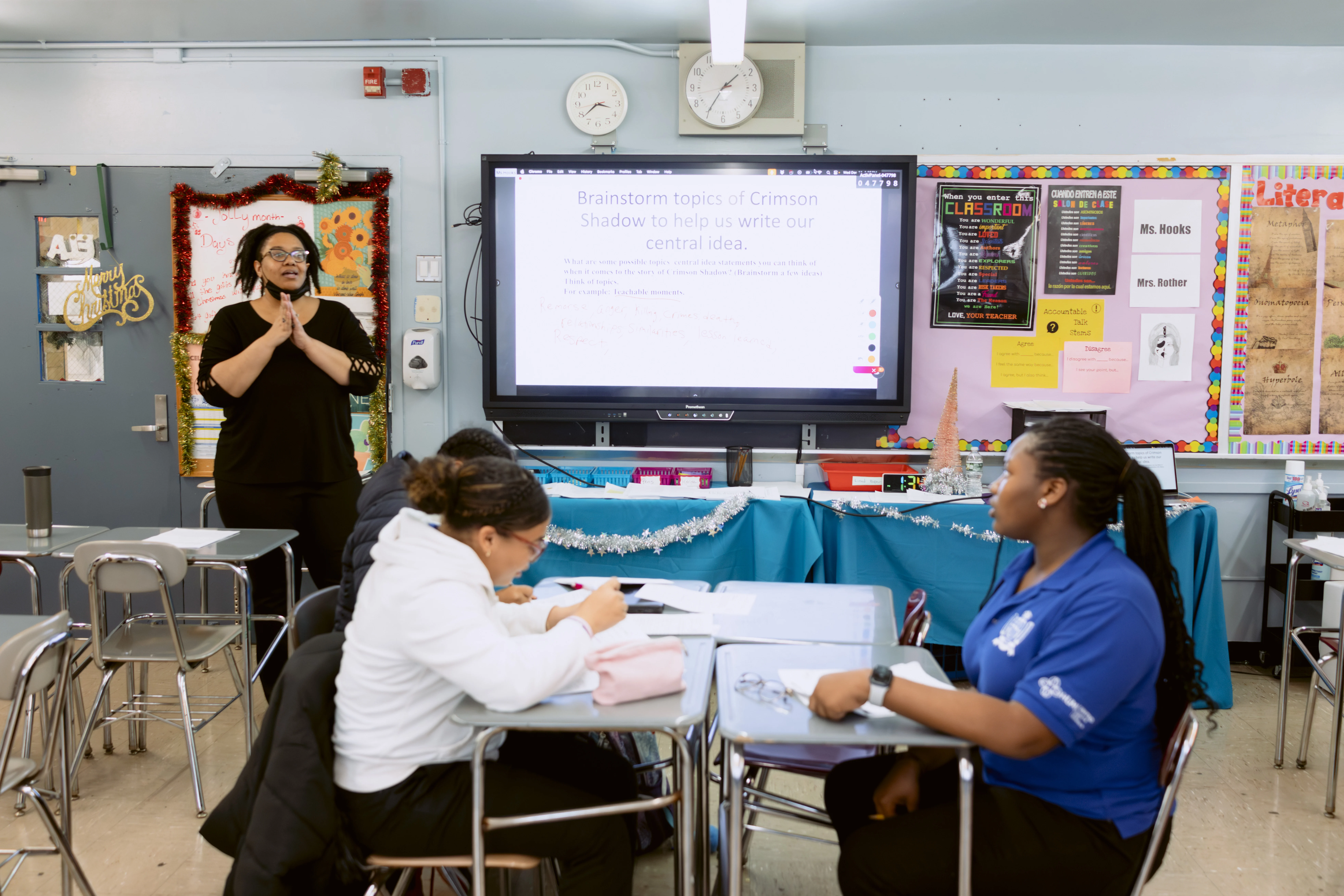 A teacher stands at the front of a classroom speaking to students seated at desks, with a brainstorming slide displayed on the interactive whiteboard.