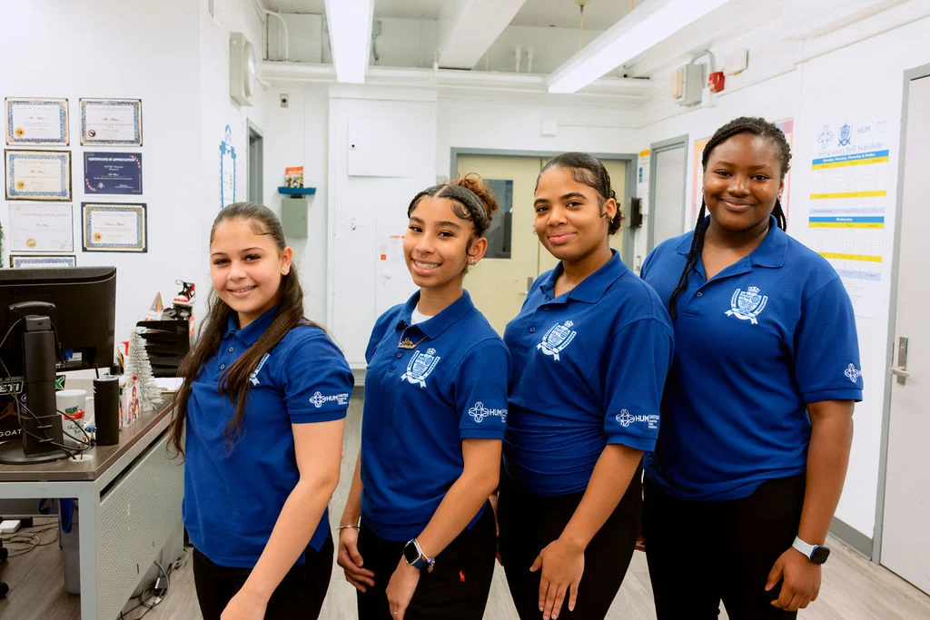 Four young women in matching blue polo shirts stand side by side in an office setting, smiling at the camera.