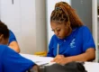 Student in blue school uniform focuses on writing at a desk, surrounded by classmates in a classroom setting.