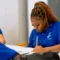 Student in blue school uniform focuses on writing at a desk, surrounded by classmates in a classroom setting.