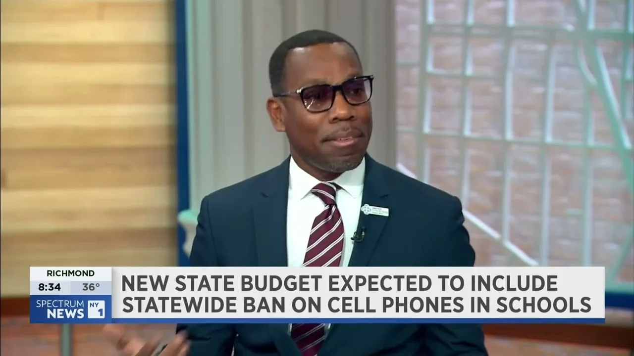 A man in a suit and glasses speaks on a news program. The screen headline discusses a possible statewide ban on cell phones in schools.
