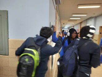 Three students with backpacks walk through a hallway. One student high-fives another who is wearing headphones. The hallway is lined with lockers and has overhead lighting.
