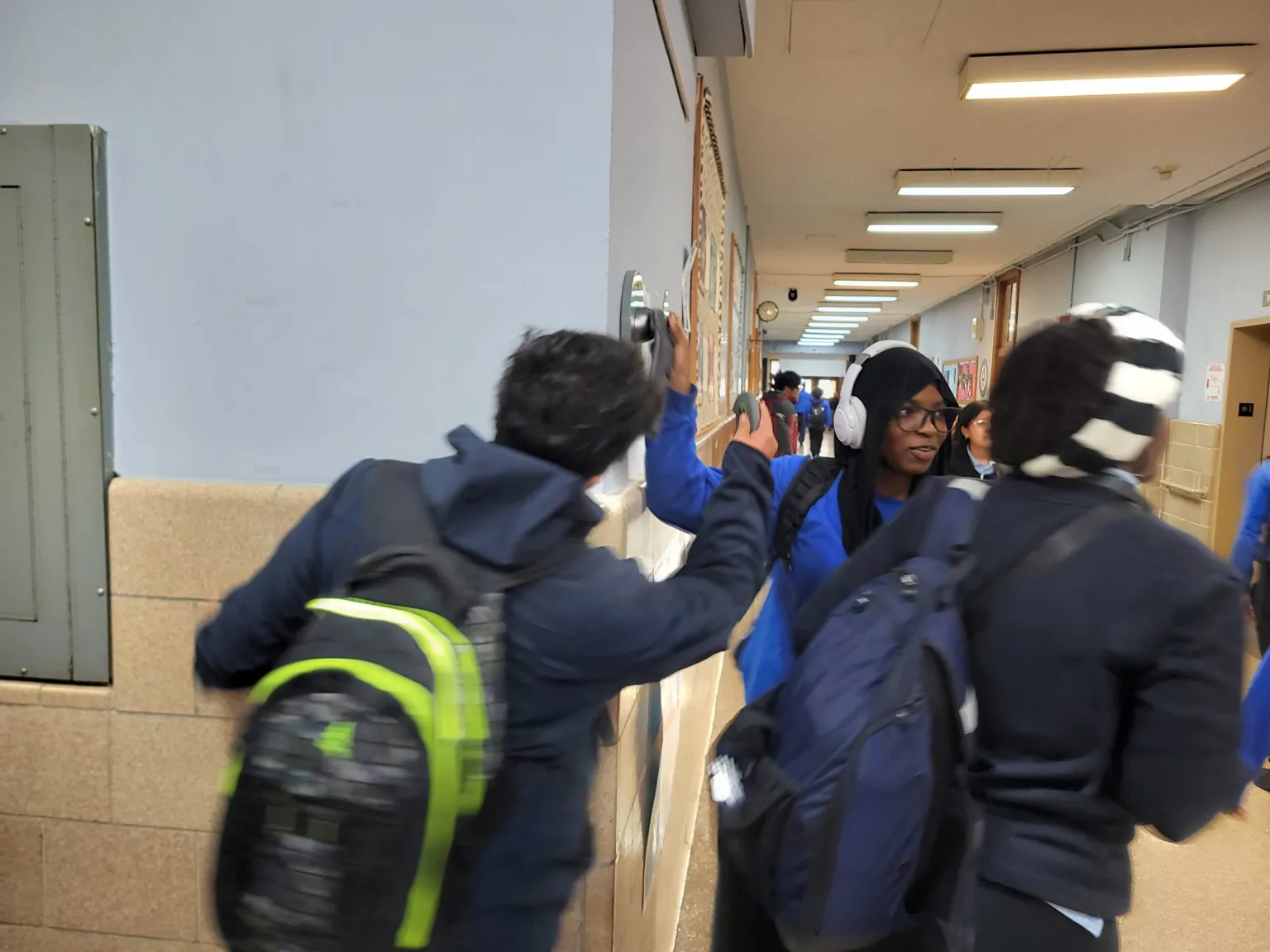 Three students with backpacks walk through a hallway. One student high-fives another who is wearing headphones. The hallway is lined with lockers and has overhead lighting.