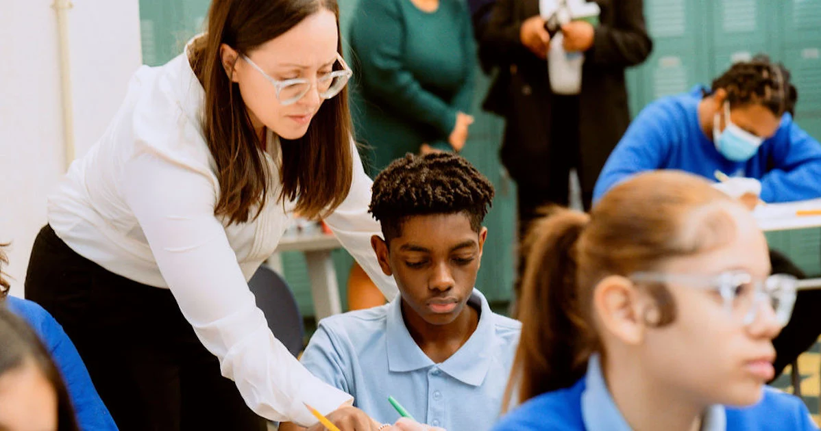 A teacher assists a student with schoolwork in a classroom. Other students write in notebooks. A person in the background wears a mask.