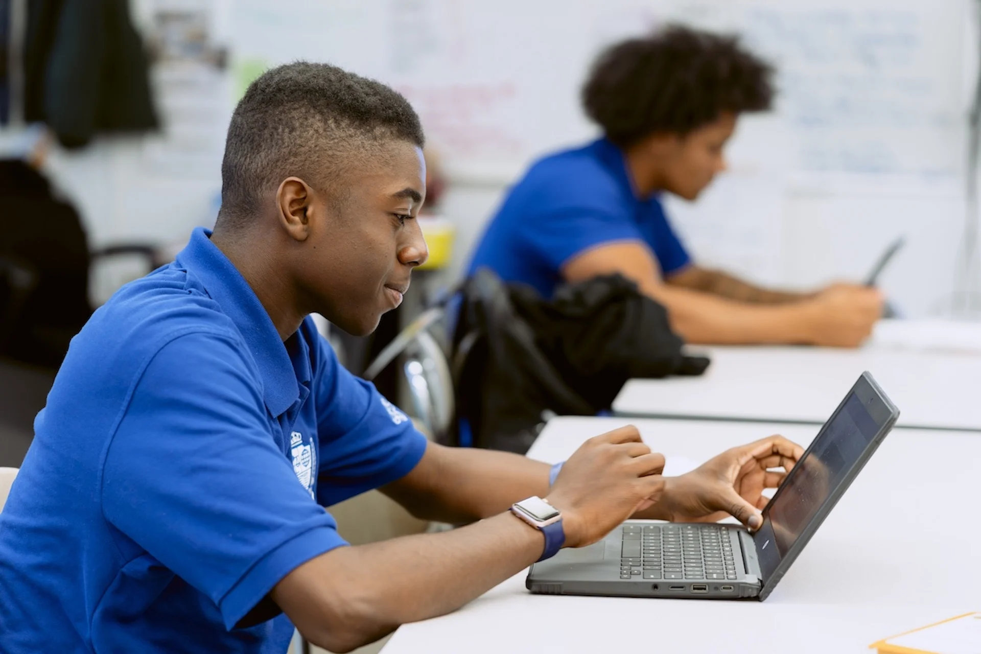 A student wearing a blue polo shirt uses a laptop at a desk in a classroom, with another student working in the background.