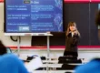 A teacher stands at the front of a classroom pointing at a screen displaying instructions while students sit at desks facing her.
