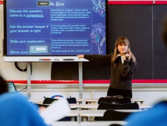 A teacher stands at the front of a classroom pointing at a screen displaying instructions while students sit at desks facing her.