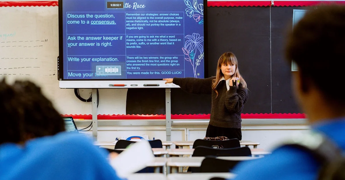 A teacher stands at the front of a classroom pointing at a screen displaying instructions while students sit at desks facing her.