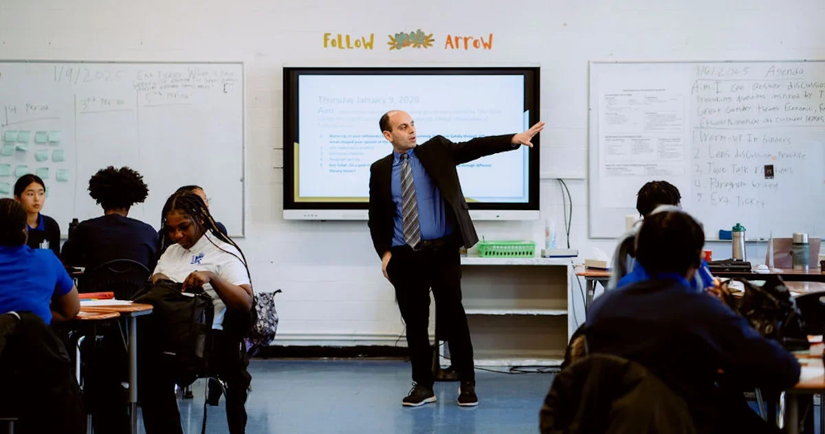 A teacher in a suit stands at the front of a classroom, pointing towards a large screen, while students sit at desks and listen.