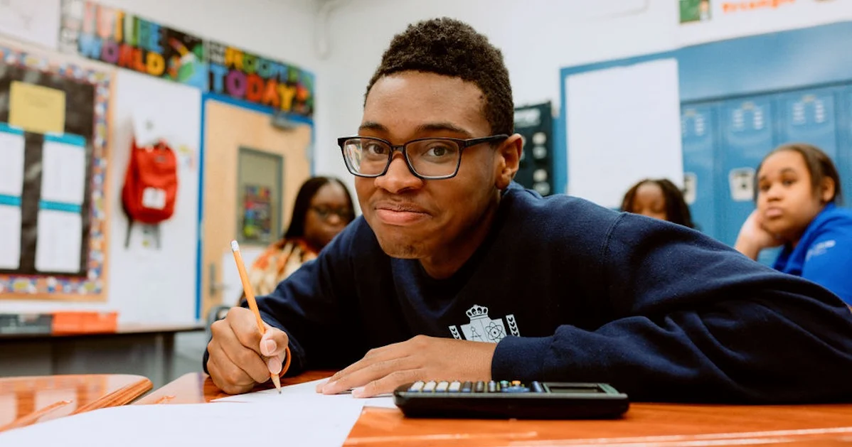  A student at United Charter Schools AMS IV smiles for a photo while sitting at a desk. He is holding a pencil and there is paper and a calculator on the desk. 