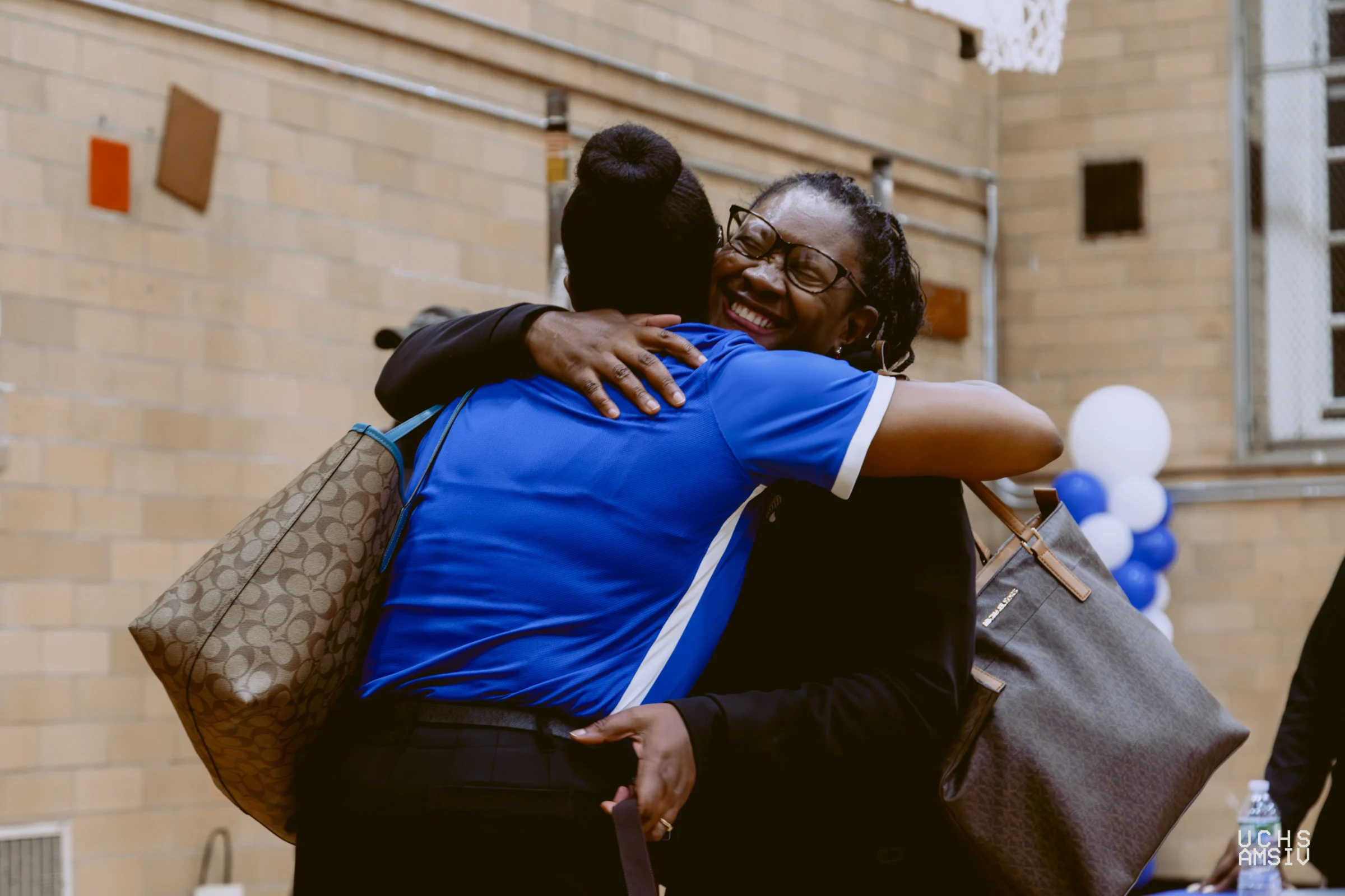 Two people hugging in a gymnasium, one wearing a blue shirt and the other in black, both carrying handbags. Blue and white balloons are visible in the background.