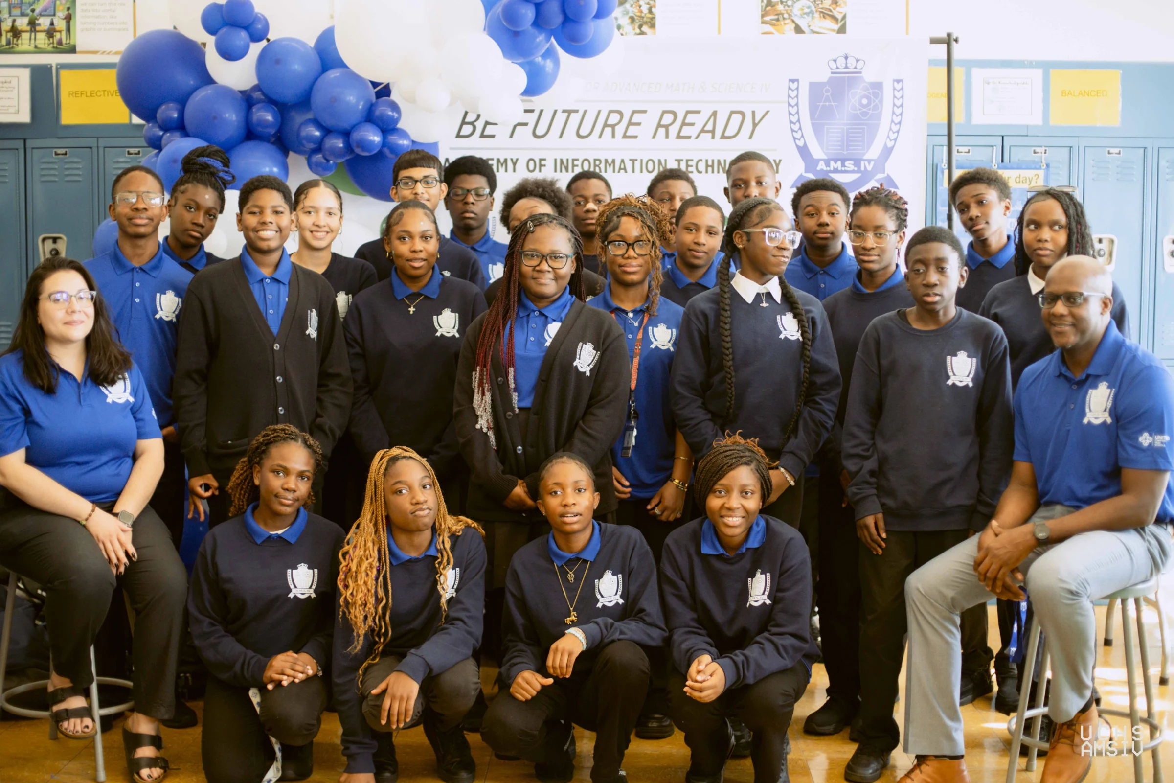 A group of students and two adults pose for a photo indoors in front of blue lockers and a banner that reads "Be Future Ready." Most wear matching blue shirts and jackets.