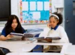 Two students United Charter High Schools HUM sit at desks in a classroom, reading textbooks. One wears a blue shirt and the other a white sweater. Classroom materials and bulletin board are visible in the background.