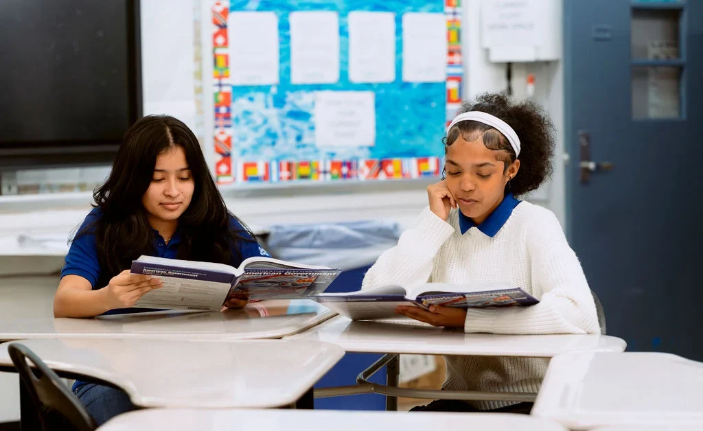 Two students United Charter High Schools HUM sit at desks in a classroom, reading textbooks. One wears a blue shirt and the other a white sweater. Classroom materials and bulletin board are visible in the background.